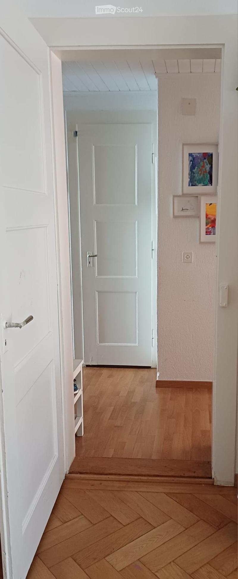 A narrow hallway with light wooden flooring and white walls, featuring a white door at the end and framed artwork on the right wall.