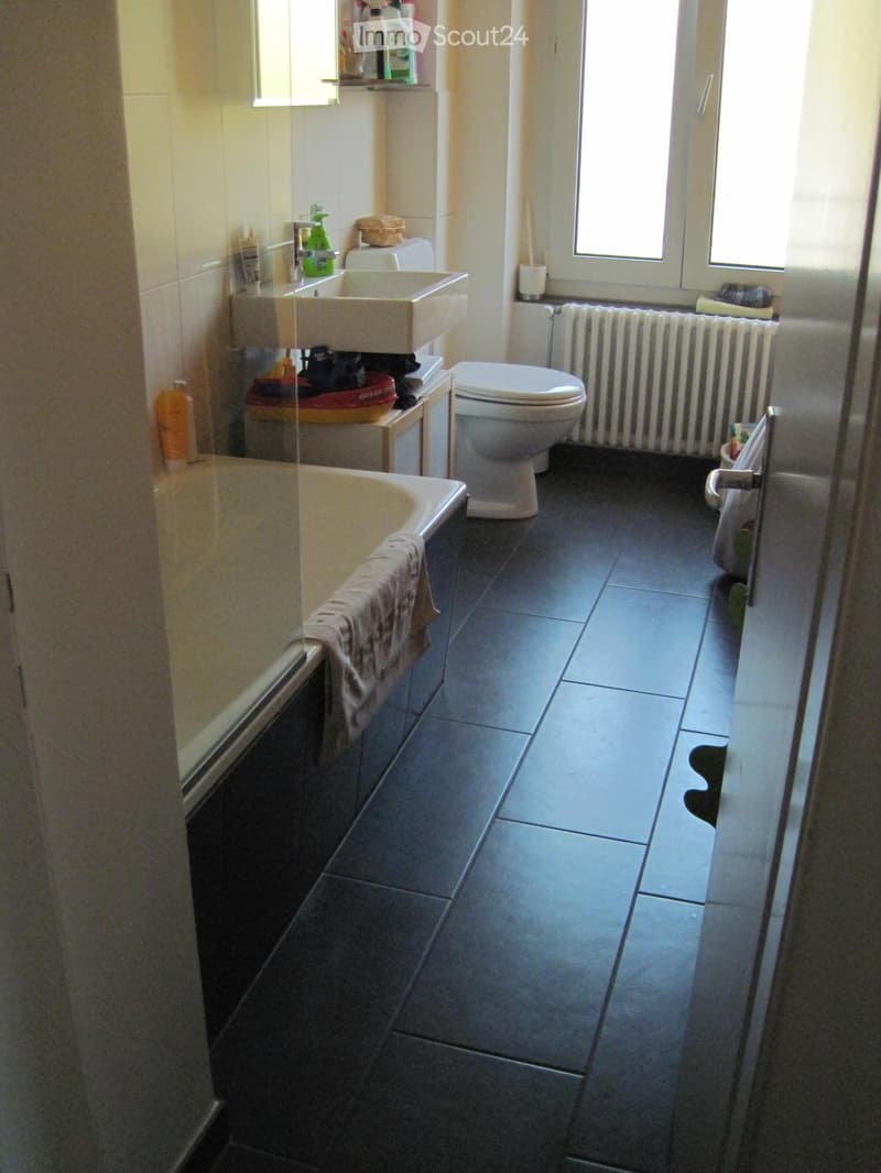 Bathroom with dark tiled floor, bathtub, toilet, sink, and radiator. Cleaning supplies and a towel are visible.