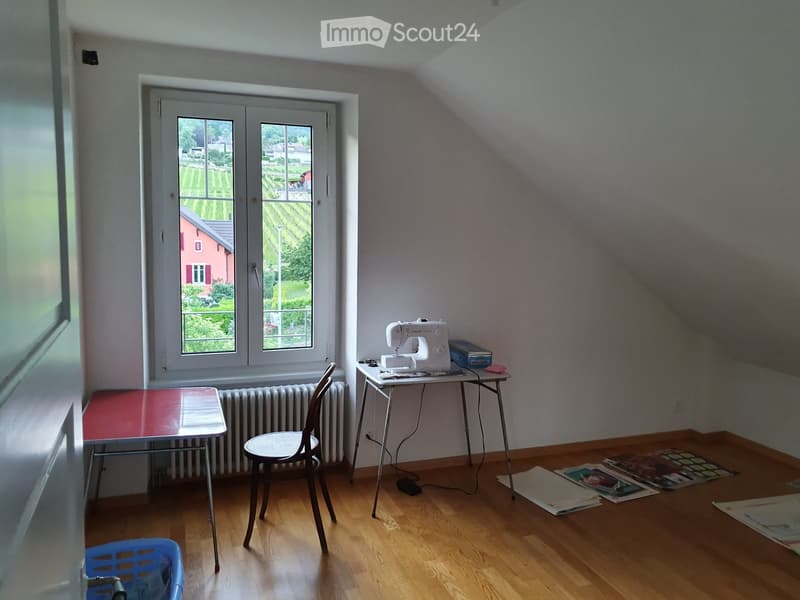 A small attic room with wooden flooring, a slanted ceiling, and large windows overlooking a vineyard. Contains a sewing machine on a table, a small table with a red top, a chair, a radiator, and a blue basket.