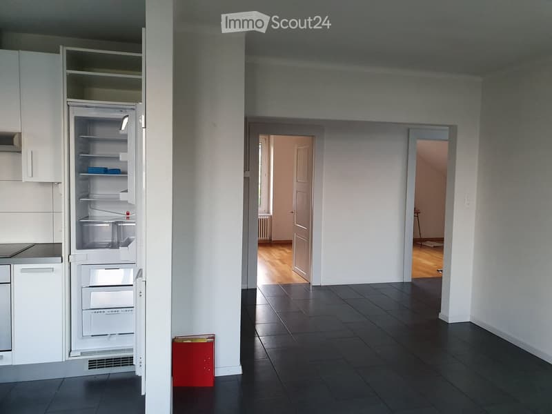 A modern, minimalist kitchen with white cabinets, a stainless steel refrigerator, and a dark tiled floor. The space is open and clean, with a red storage box visible near the entrance.