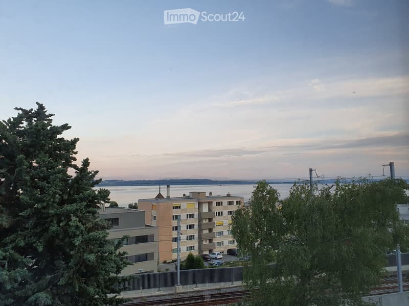 Multi-story residential building with balconies and windows, surrounded by trees and a body of water in the background.