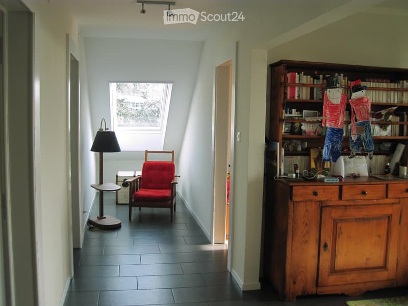 A cozy, well-lit room with a slanted ceiling and skylight. Features include a wooden chair with a red seat, a small round table with a lamp, a large wooden bookcase filled with books and decorative items, and a wooden cabinet with drawers.