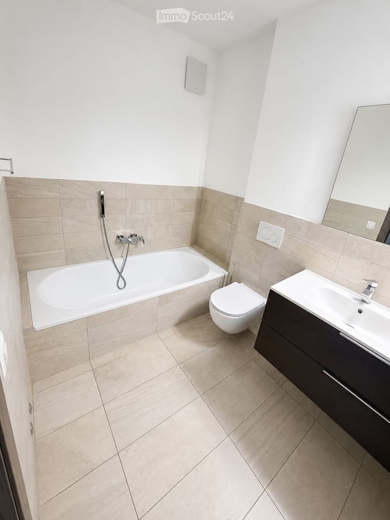 Modern bathroom with a bathtub, toilet, sink, and mirror. Beige tiles on walls and floor, sleek dark vanity cabinet, and modern fixtures.