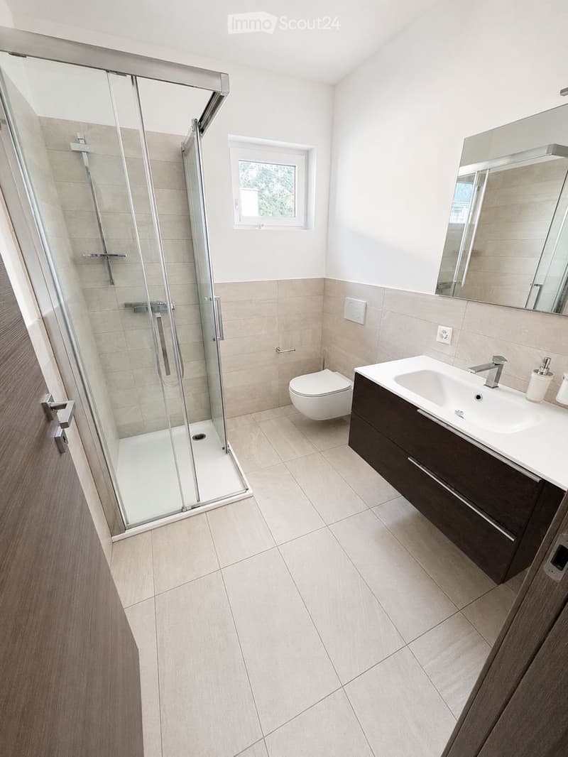 Modern bathroom with a white tiled shower enclosure, a white sink with a dark wooden cabinet, a toilet, and a large mirror above the sink. The floor is tiled in light beige tiles.