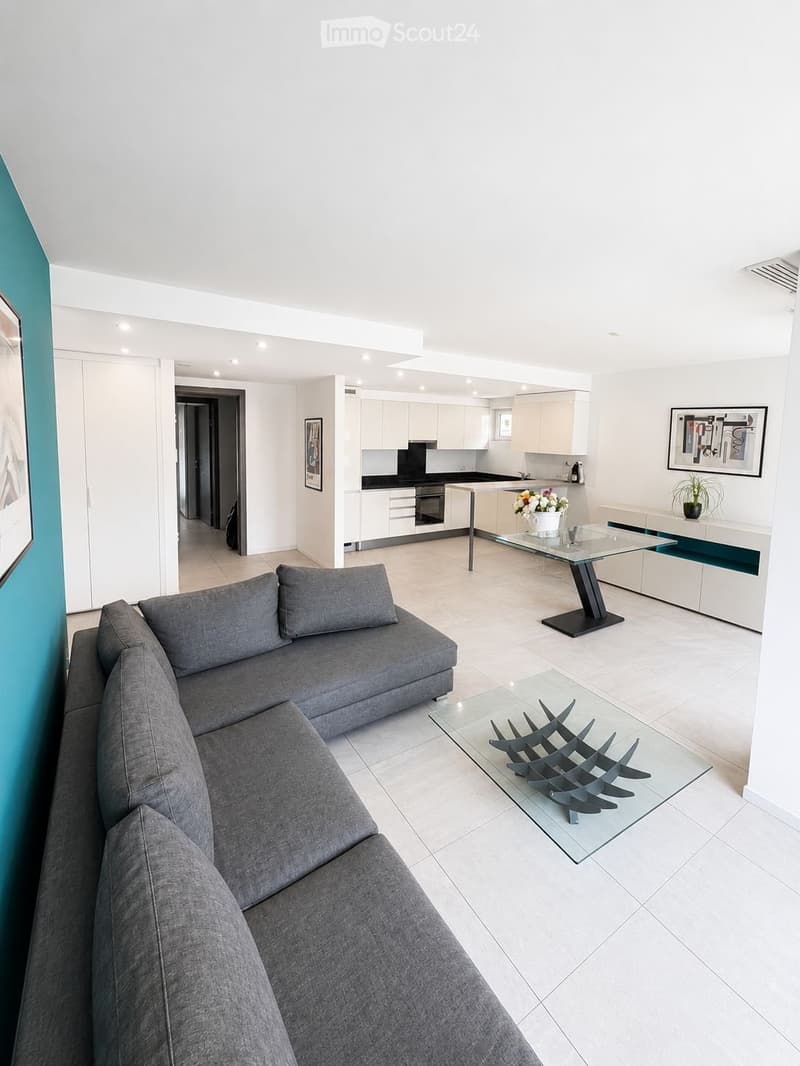 Modern living room with a grey sectional sofa, glass coffee table, and a sleek kitchen area with white cabinets and stainless steel appliances. The space features a white tiled floor, a glass dining table, and decorative artwork on the walls.