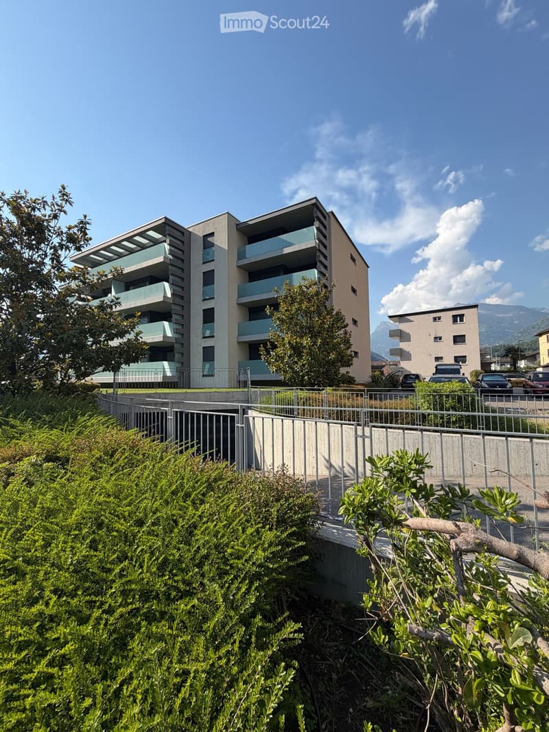 Modern multi-story residential building with balconies, surrounded by greenery and a raised ground floor entrance.