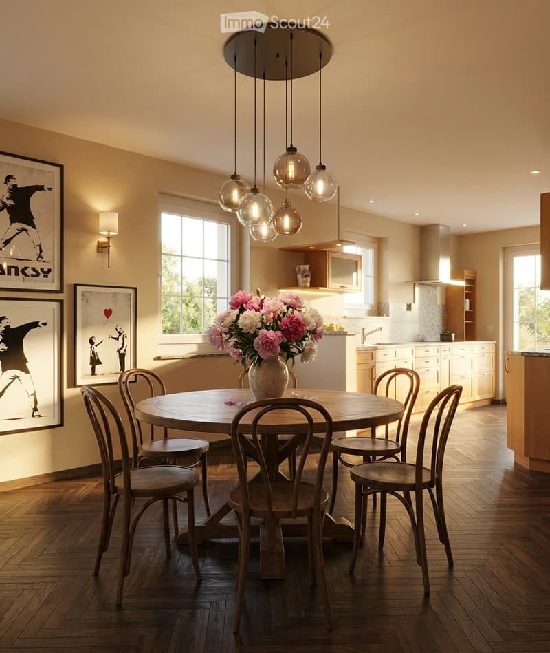 Open-plan dining and kitchen area with a round wooden table surrounded by six wooden chairs, a vase of pink and white flowers, framed artwork on the walls, and a modern pendant light fixture above the table. The kitchen features white cabinets, a sink, an