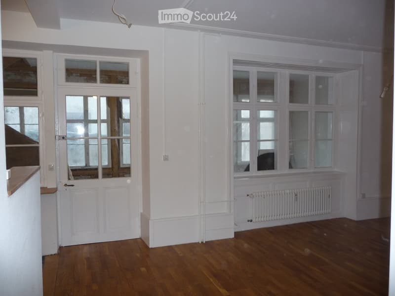 Empty room with wooden flooring, white walls, large windows, and a radiator. No furniture or equipment present.