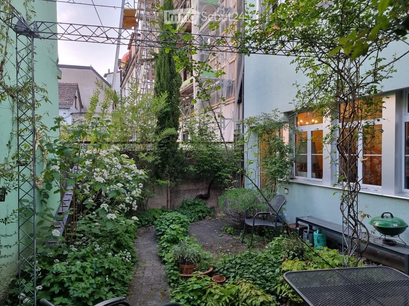 A lush, narrow garden with climbing plants, potted plants, a metal table, chairs, a bench, and a grill, surrounded by buildings.