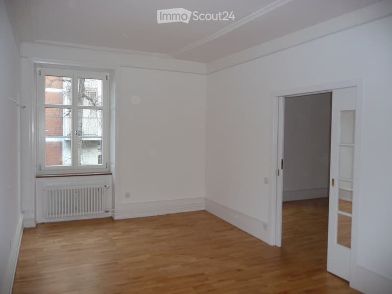 Empty room with wooden flooring, white walls, a window with white frame, radiator, and an open doorway leading to another space.