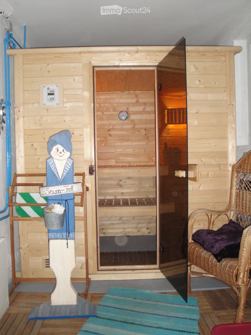 A wooden sauna room with a control panel, thermometer, a decorative statue, a wicker chair with a pillow, and a blue rug.