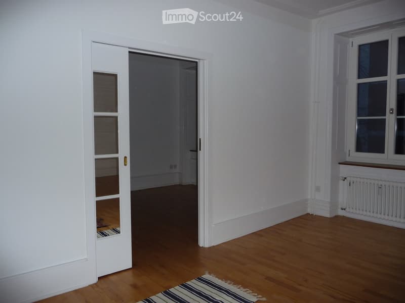 Empty room with wooden flooring, white walls, and a partially open door leading to another room. A small rug is placed near the door.