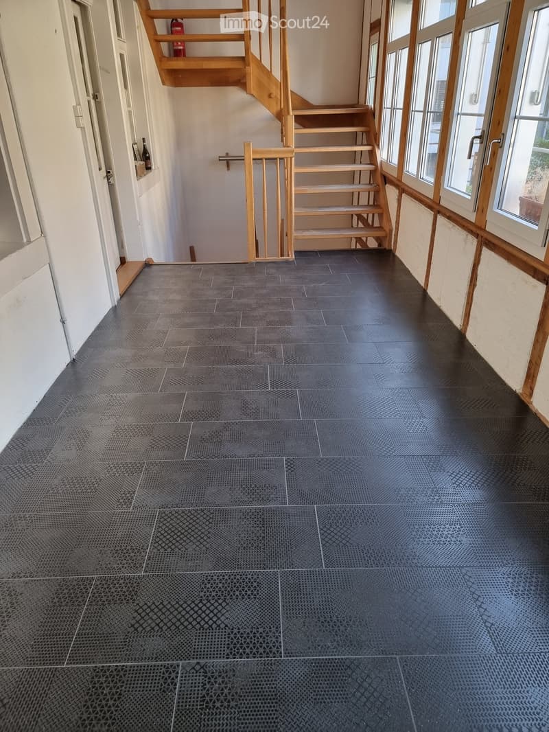 Empty hallway with dark grey tiled floor, wooden staircase, and large windows on the right side.
