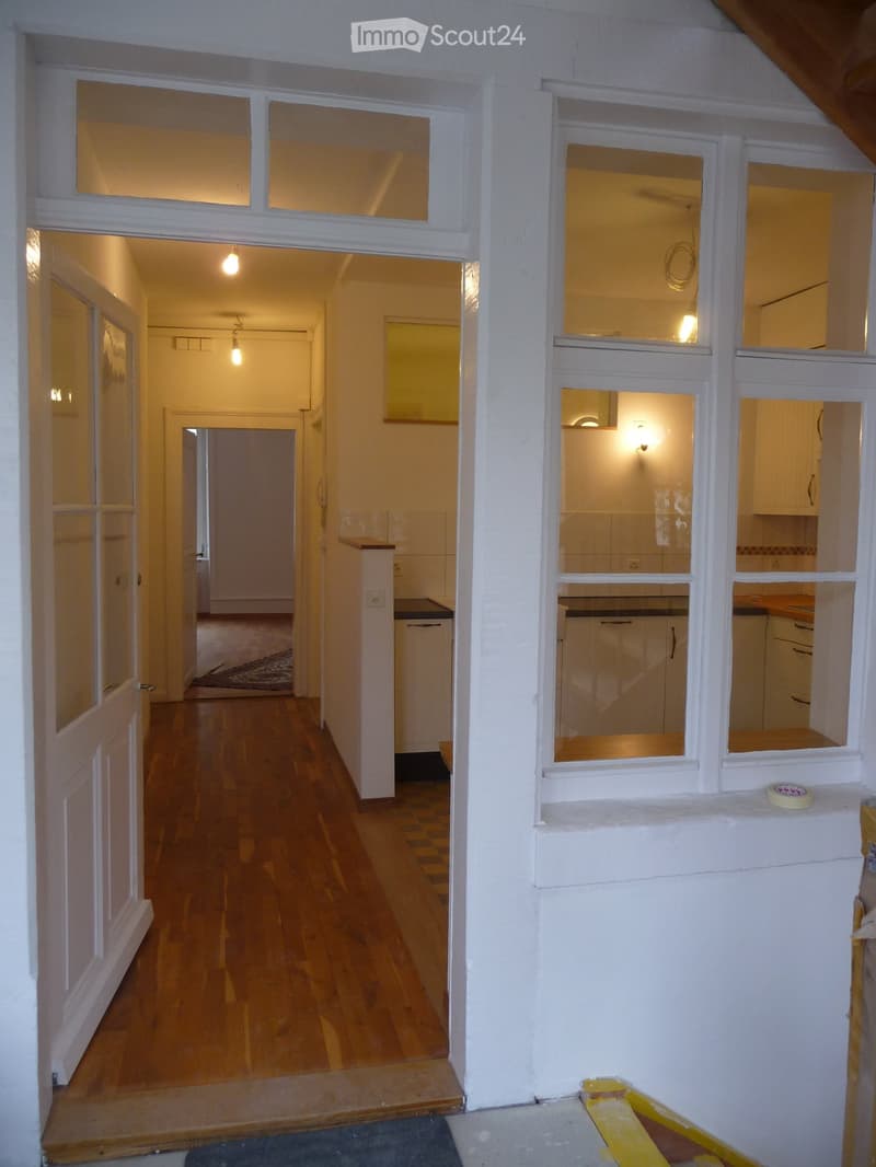 A well-lit hallway with wooden flooring, white walls, and multiple doors leading to different rooms, including a kitchen and a living area.