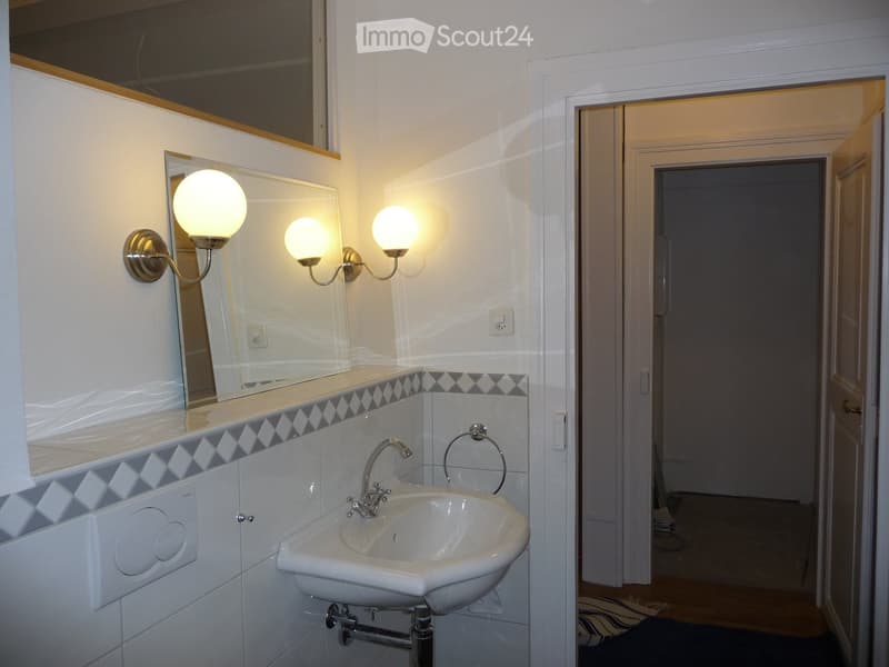 Bathroom with a white sink, mirror, and wall-mounted light fixtures. The walls are tiled with a decorative border, and a door leads to another room.