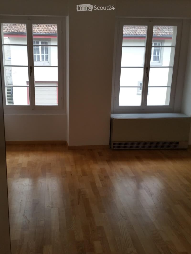 Empty room with wooden flooring, large windows, and radiator on the wall.