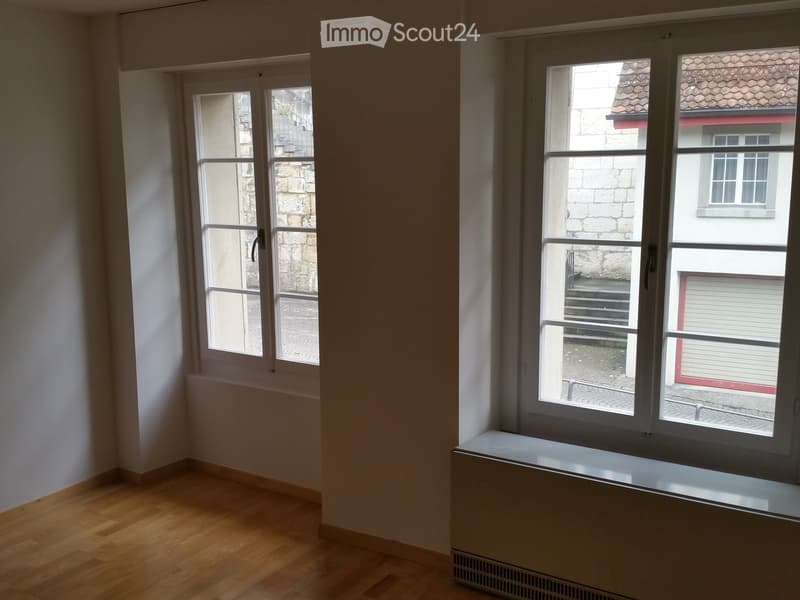Empty room with wooden flooring, white walls, and large windows offering views of a stone building and a tiled roof.