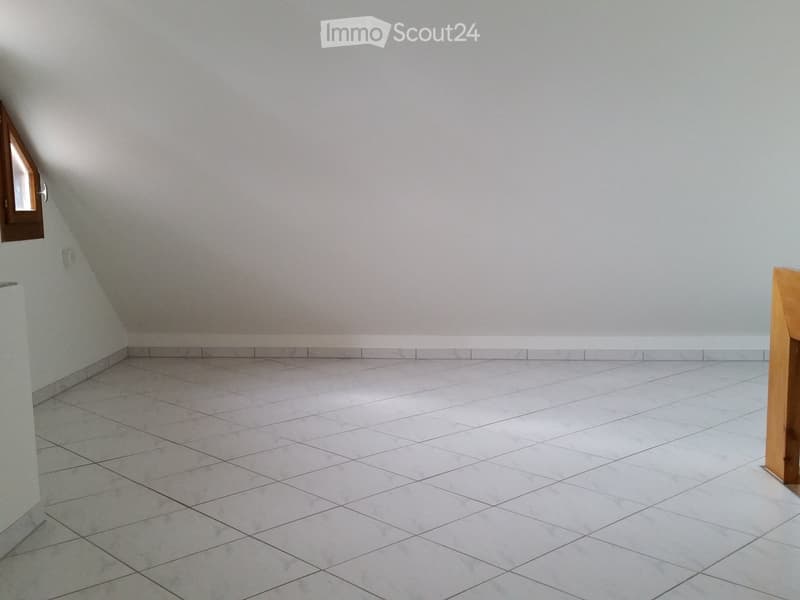 Empty room with light grey walls and white tiled floor, featuring a small window on the left side and a wooden door frame on the right.