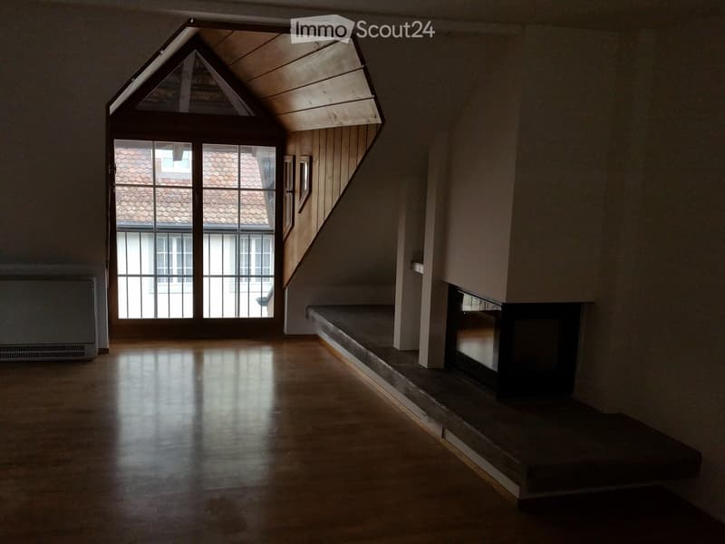 Empty room with wooden floor, large window, fireplace, and slanted wooden ceiling.