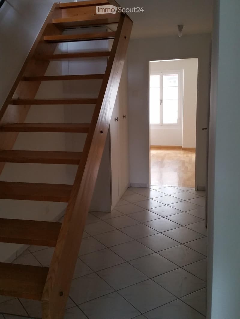 Empty hallway with tiled floor, wooden staircase leading upwards, and a doorway leading to another room with a window.