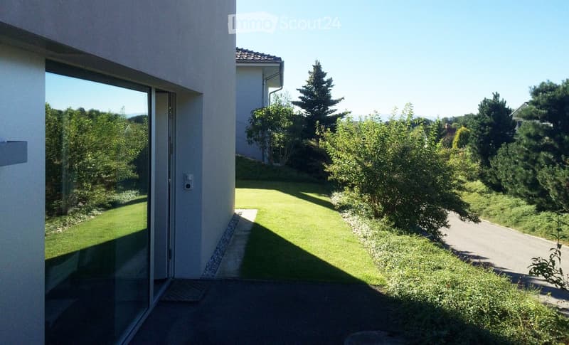 View from a modern house's balcony or patio showing a well-maintained lawn, trees, and a pathway leading to the house.