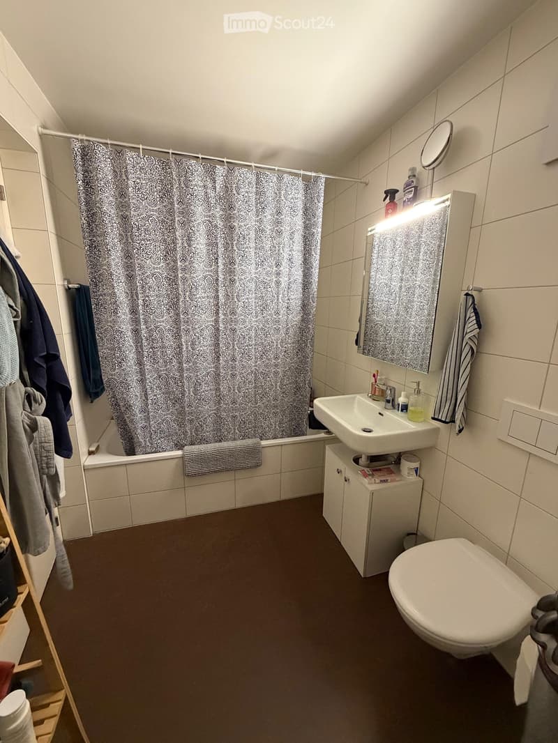 Modern bathroom with a bathtub, shower curtain, sink, mirror, toilet, and cabinet. Towels and toiletries are visible.