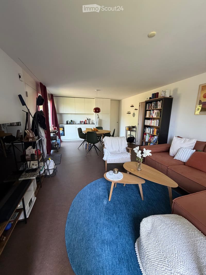 Modern living room with a blue round rug, a brown sofa, a white armchair, wooden coffee tables, a bookshelf, and a dining area with a round table and chairs. The room features white walls, wooden flooring, and a kitchen area with white cabinets.