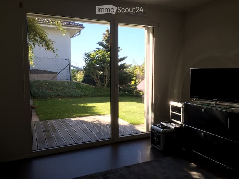 Bright living room with large sliding glass doors opening to a wooden deck and well-maintained garden. Features a flat-screen TV on a sleek black cabinet and a small black case on the floor.
