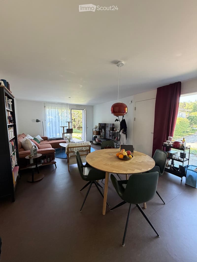 Modern living and dining area with a round wooden dining table, green chairs, a pink sofa, a bookshelf, a cat tree, and a television. The space features large windows with curtains and a view of greenery outside.