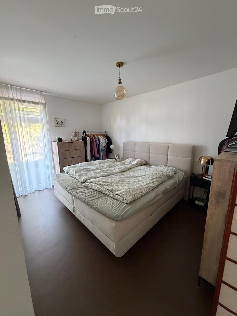 A well-lit bedroom with a large bed, nightstand, lamp, and a clothing rack with hanging clothes. The room features a wooden floor and a window with sheer curtains.