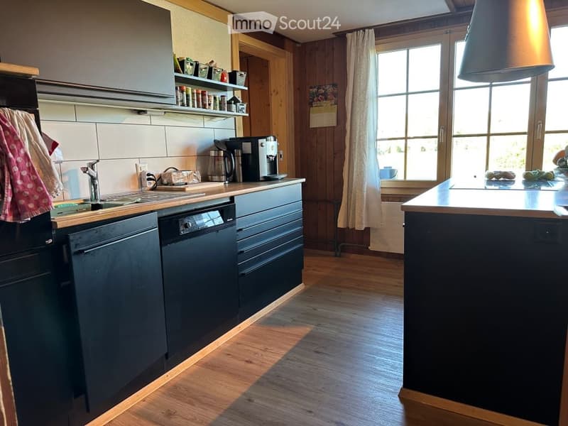 Modern kitchen with wooden flooring, black cabinets, stainless steel appliances, and a large island. Features include a sink with a faucet, a coffee maker, a dishwasher, and a window with curtains.
