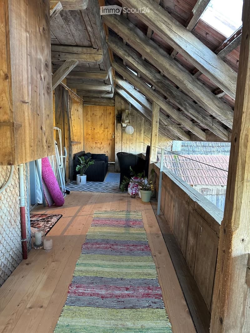 A cozy, wooden-floored attic space with exposed wooden beams, a colorful runner rug, a sofa, a chair, potted plants, and candles.