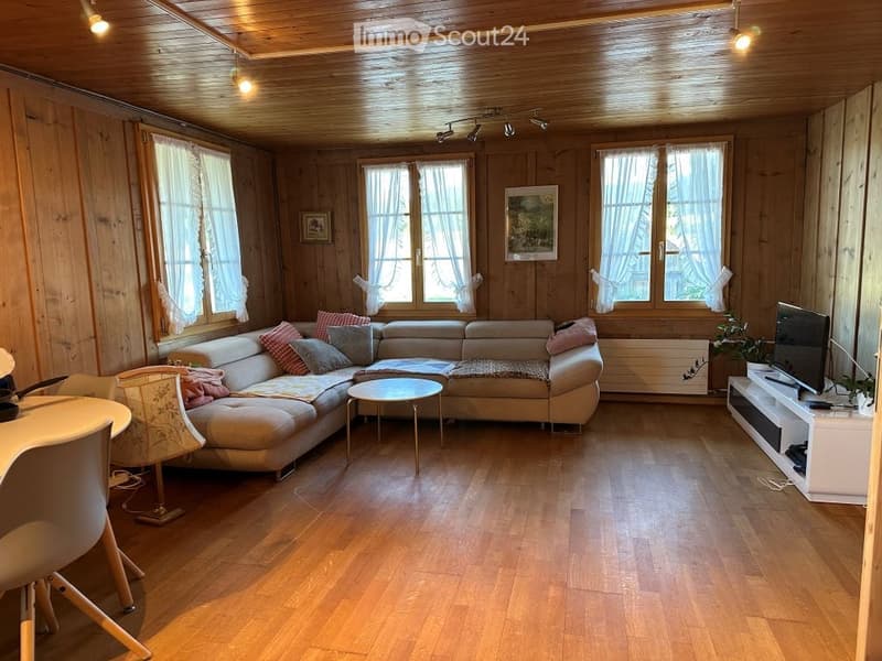 Cozy living room with wooden walls and ceiling, large windows with white curtains, a beige sectional sofa with colorful pillows, a small round coffee table, a white TV stand, and a dining area with a table and chairs.