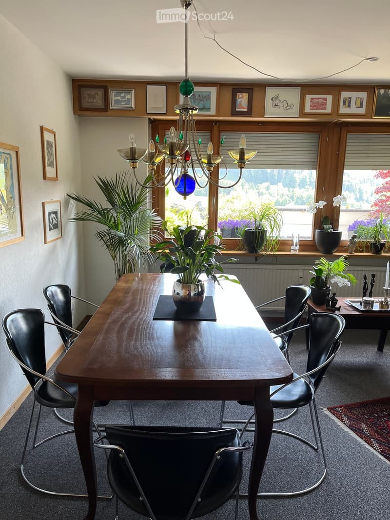 Dining room with a wooden table, black chairs, chandelier, and potted plants on the windowsill.