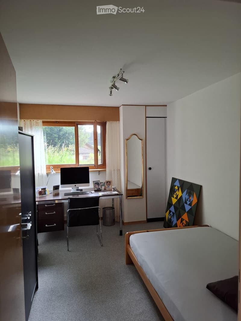 Well-lit bedroom with a bed, desk, chair, computer, mirror, and artwork. Large windows with curtains provide natural light.