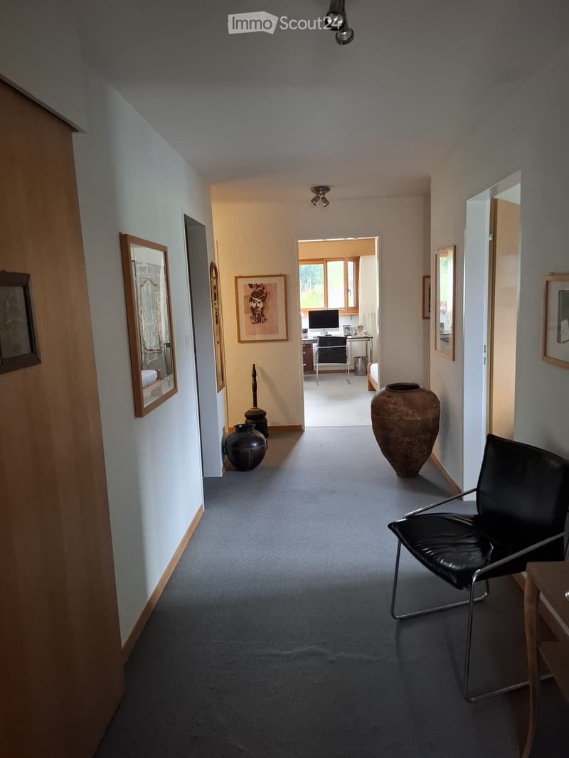 A well-lit hallway with grey carpeting, white walls, and several framed artworks. A black leather chair is positioned on the right, and large ceramic vases are placed along the left wall. The hallway leads to an open area with a desk and computer visible