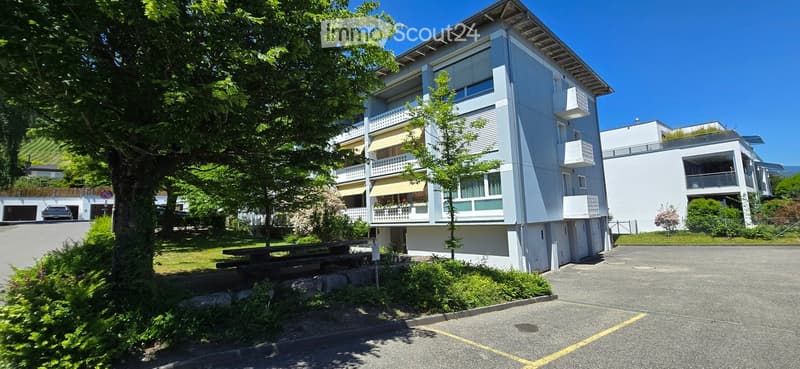 Modern multi-story apartment building with balconies, parking area, and surrounding greenery.