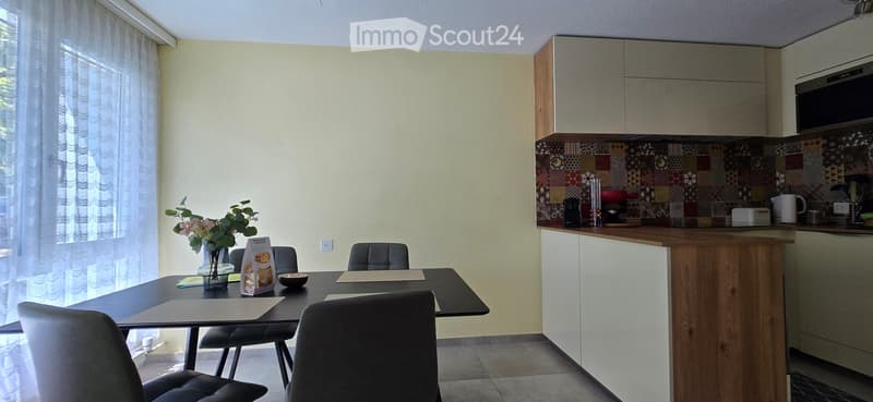 Modern kitchen with a dining area, featuring a wooden island, grey chairs, a vase with flowers, and various kitchen appliances.
