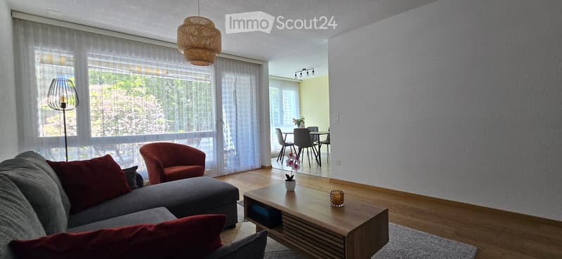 Modern living room with a grey sectional sofa, red cushions, a wooden coffee table, a red armchair, and a floor lamp. The room features large windows with sheer curtains and a view of greenery outside. A dining area with a table and chairs is visible in t
