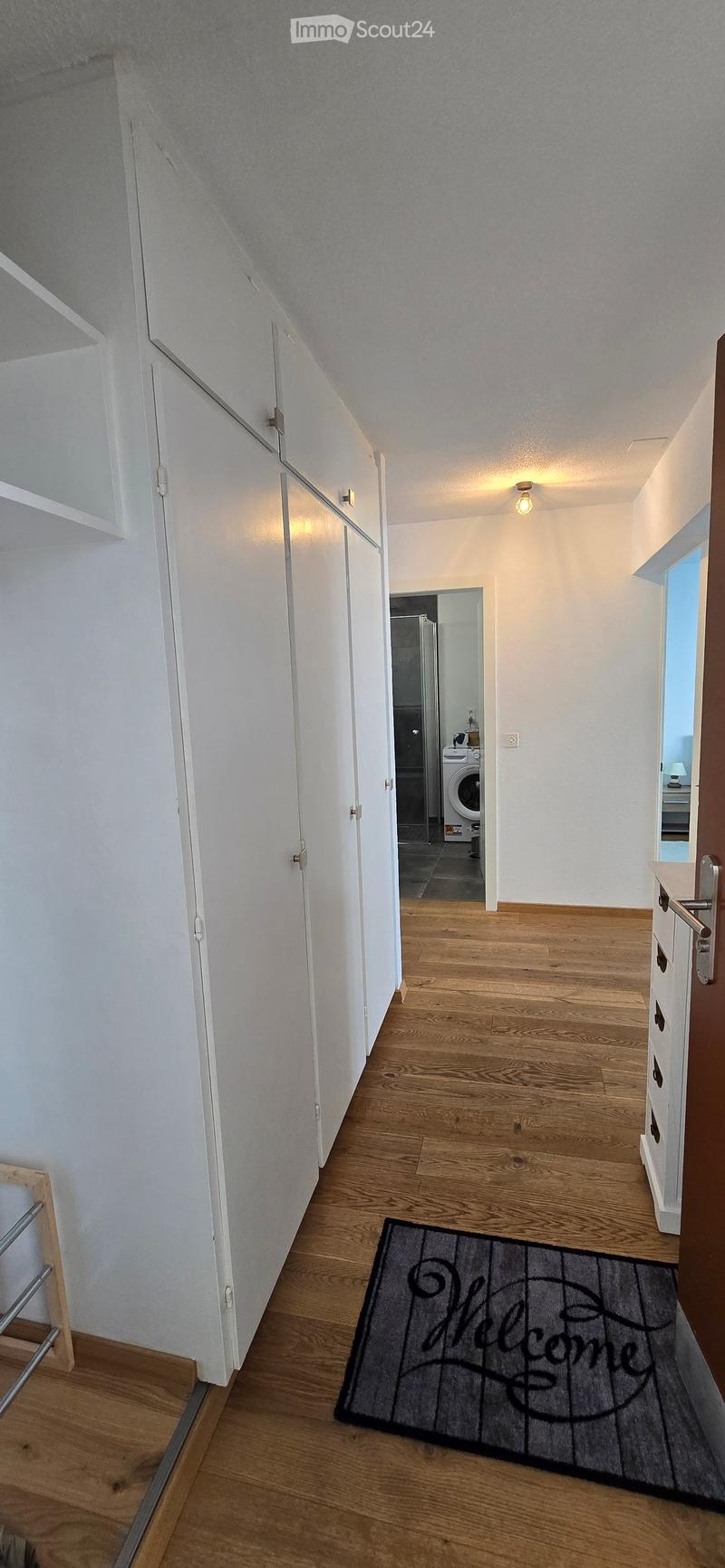 A well-lit hallway with wooden flooring, white walls, and built-in storage cabinets. A welcome mat is placed on the floor, and a washing machine is visible in an adjacent room.