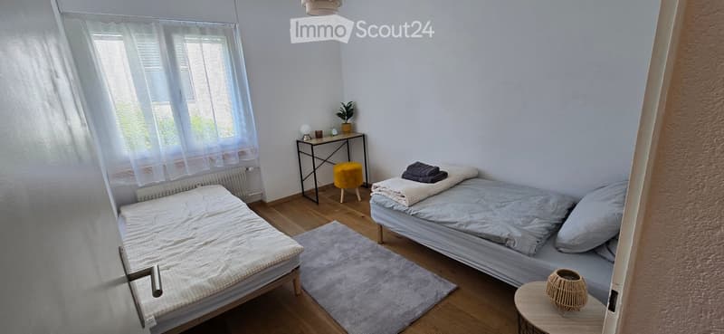 Cozy bedroom with two single beds, a small table with a lamp, a yellow stool, and a plant on the windowsill.