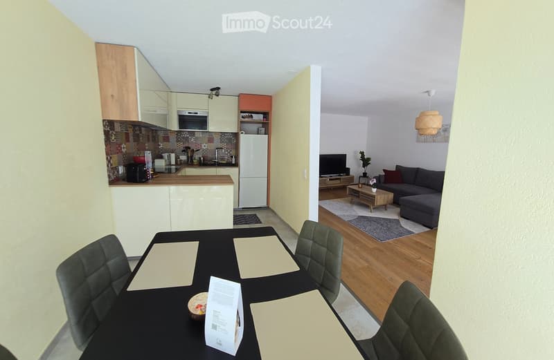 Open-plan living and dining area with modern furniture, including a sofa, coffee table, dining table with chairs, and a kitchen area with cabinets, refrigerator, and oven.