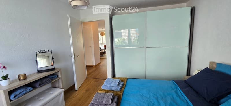 Modern bedroom with a large bed, blue bedding, a tall wardrobe with mirrored doors, a nightstand with a mirror, and a small shelf holding towels and a flower pot.