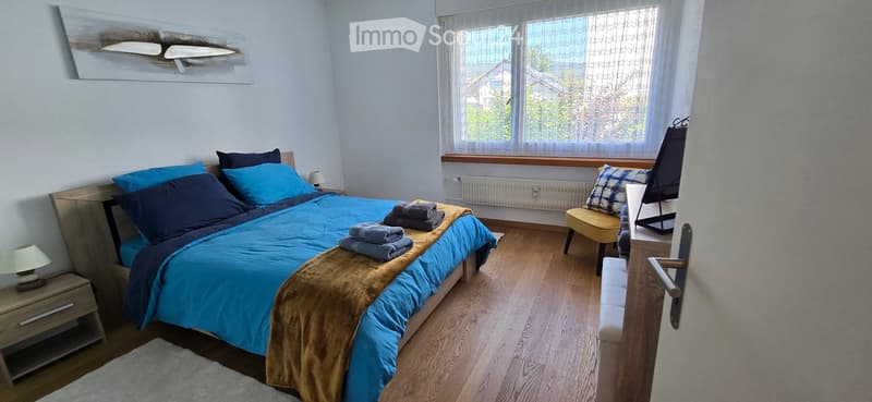 Cozy bedroom with a wooden bed, blue bedding, nightstand, chair, and a mirror. Natural light from a window with sheer curtains.