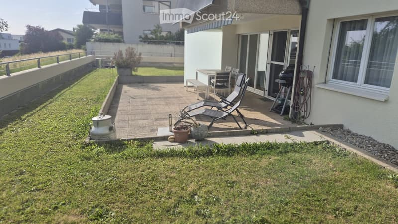 Garden area with a paved patio, lounge chairs, a table and chairs, barbecue, and potted plants. The lawn is bordered by a low concrete wall and a railing.