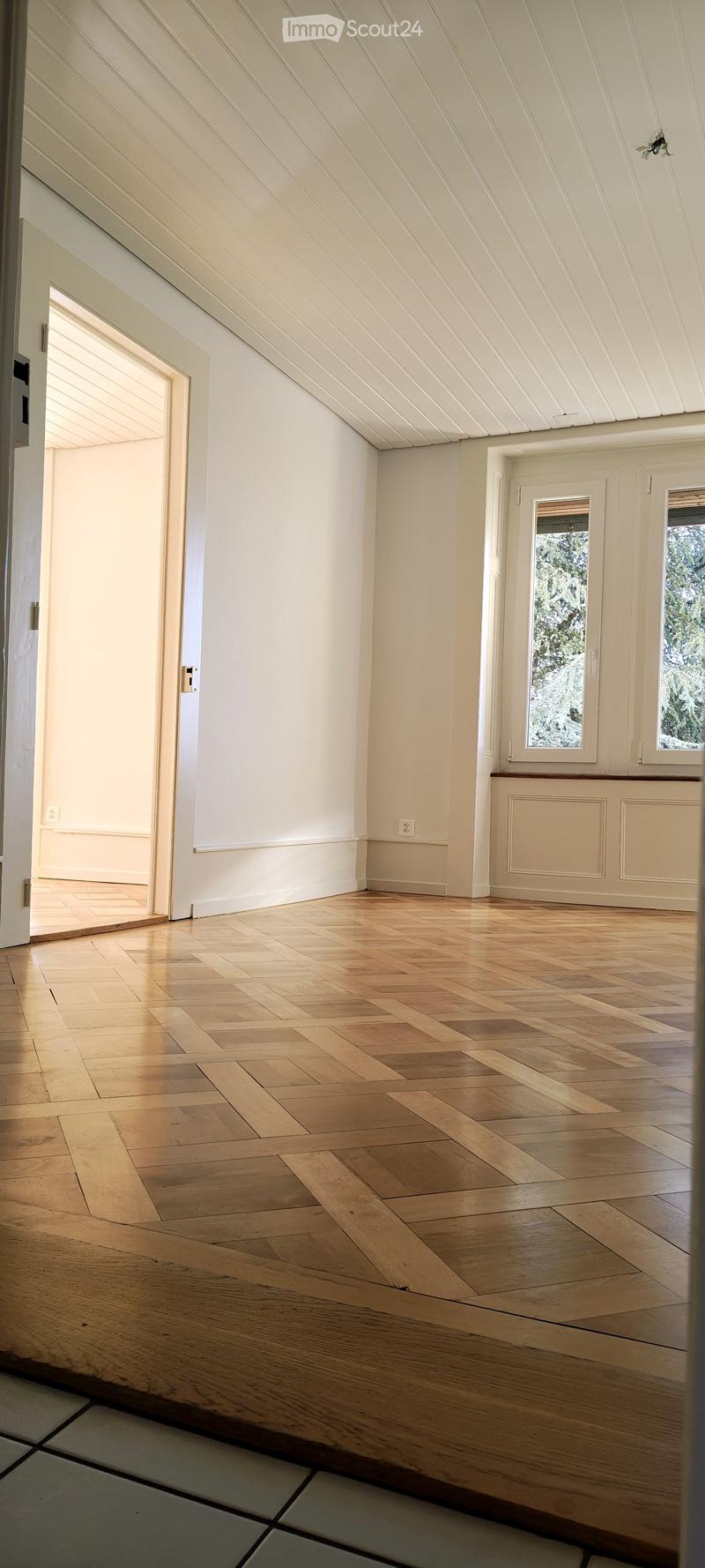 Empty room with wooden herringbone flooring, white walls, and large windows showing outdoor view. No furniture or equipment present.