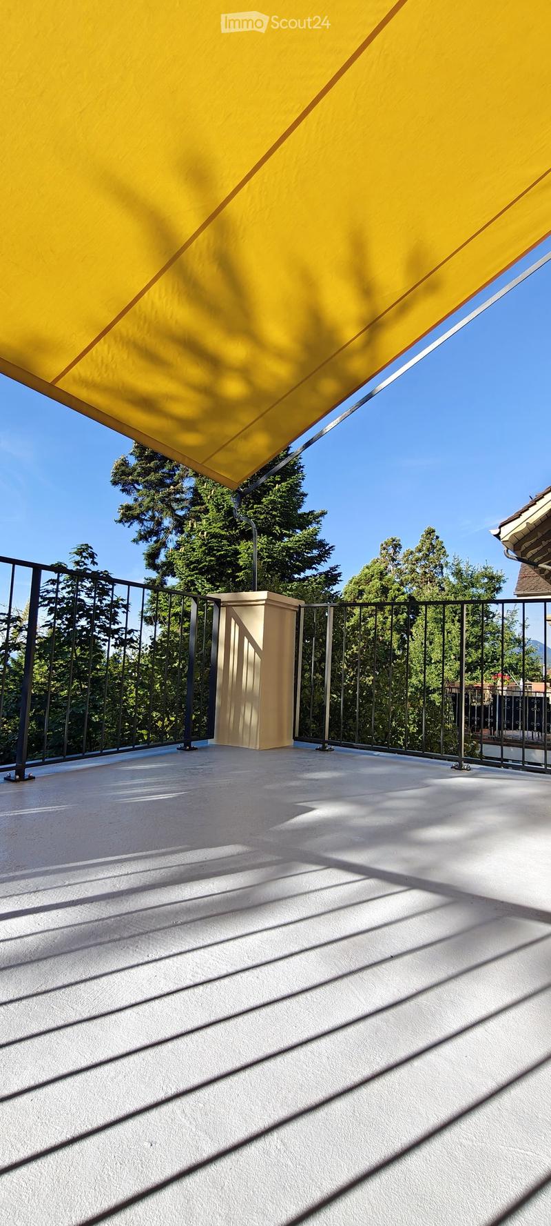 Balcony with a yellow awning, metal railing, and a small beige structure, surrounded by trees and a clear blue sky.