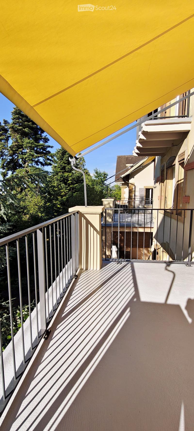 Balcony with metal railing, yellow awning, and view of trees and neighboring building.