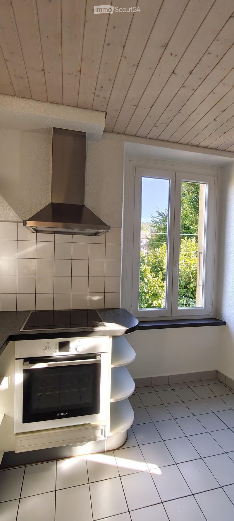 Modern kitchen with white tiled walls, white tiled floor, Bosch oven, range hood, and induction cooktop. Large window with a view of greenery.