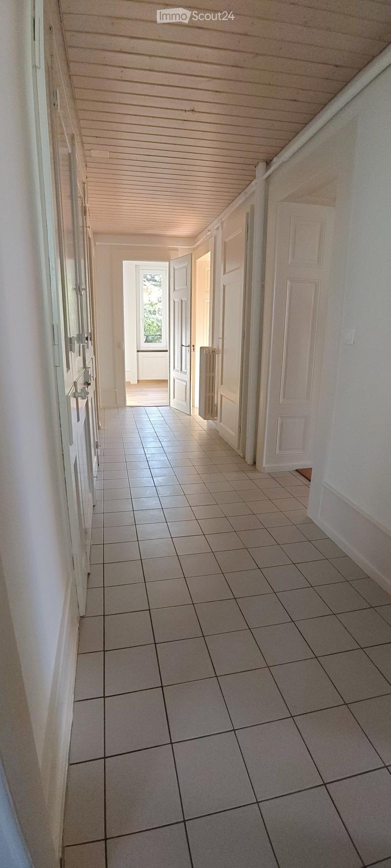 A clean, tiled hallway with white walls, multiple doors, and a slanted wooden ceiling. Natural light streams in from an open doorway at the end.